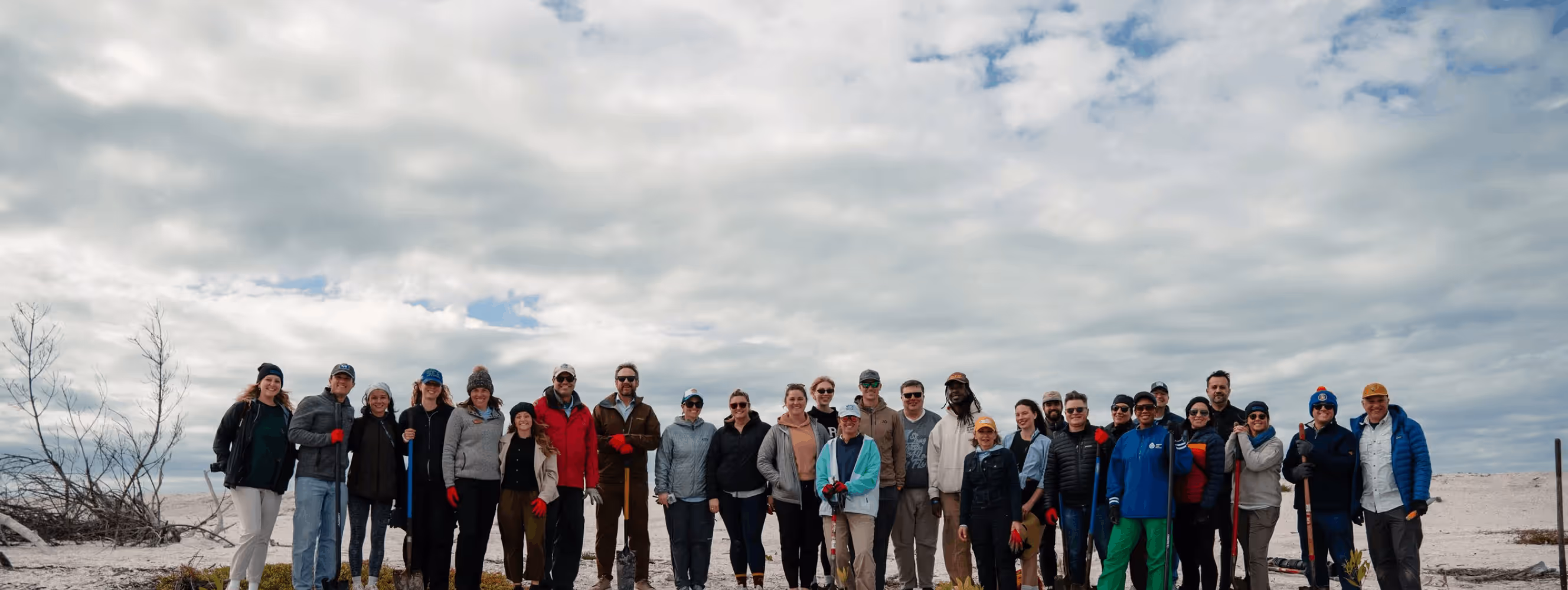 Group of people standing side by side outdoors on a cloudy day, many holding gardening tools.