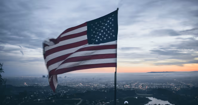 American flag waving on a pole overlooking a cityscape at sunset with cloudy skies.