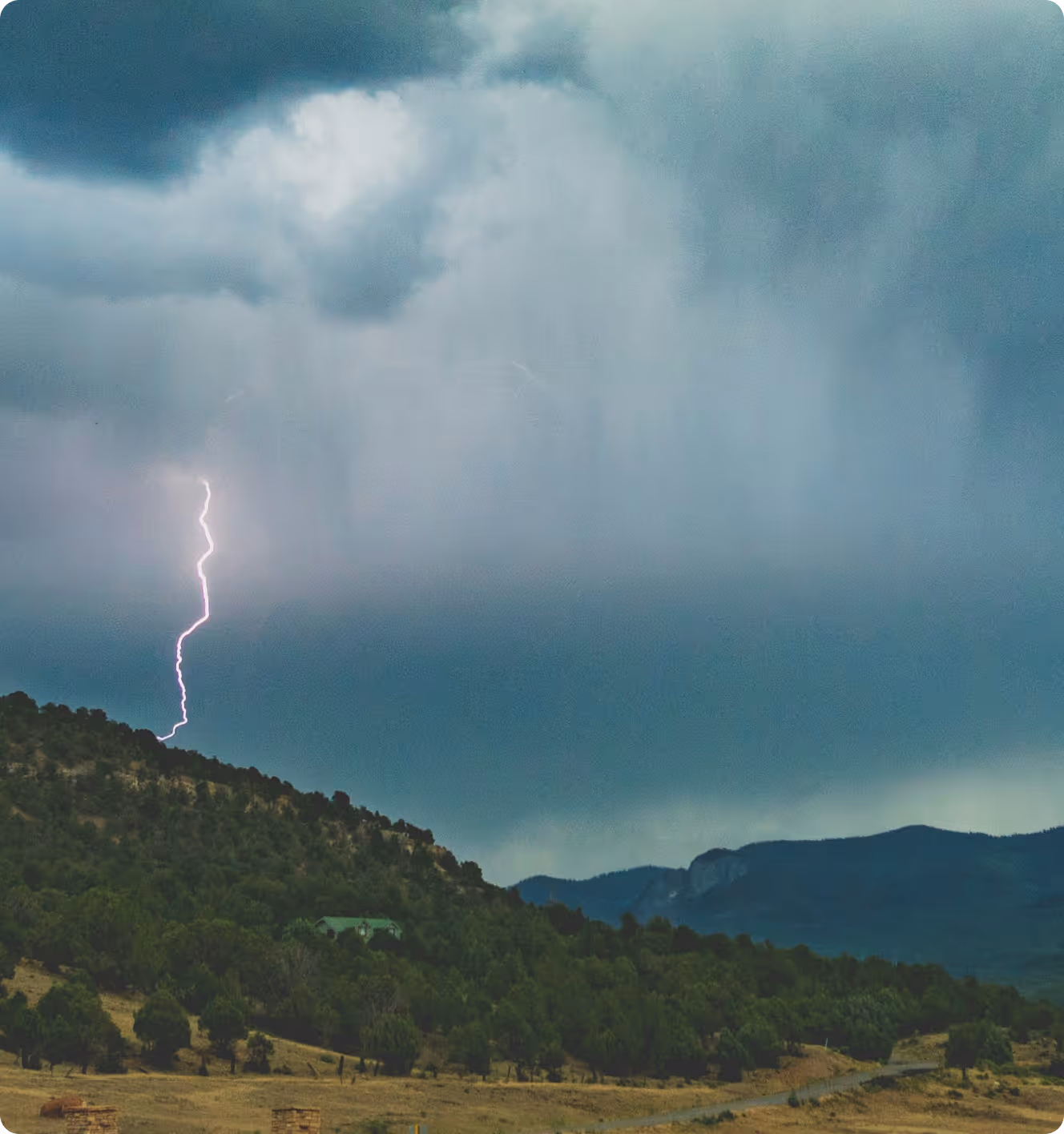 Lightning striking a forested hillside with dark storm clouds overhead and mountains in the background.