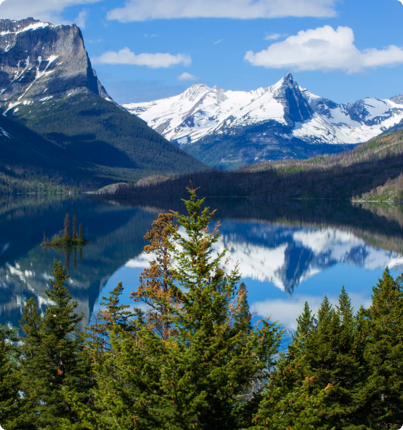 Snow-capped mountains reflecting in a clear blue lake surrounded by pine trees under a partly cloudy sky.