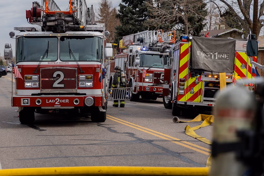Two red and white fire trucks with ladders on a street, firefighters and coiled yellow hose visible.