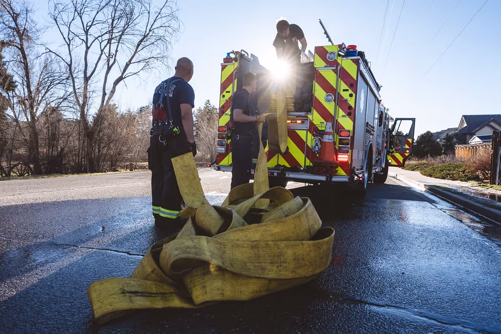Firefighters handling a large yellow fire hose near the back of a fire truck on a wet road.