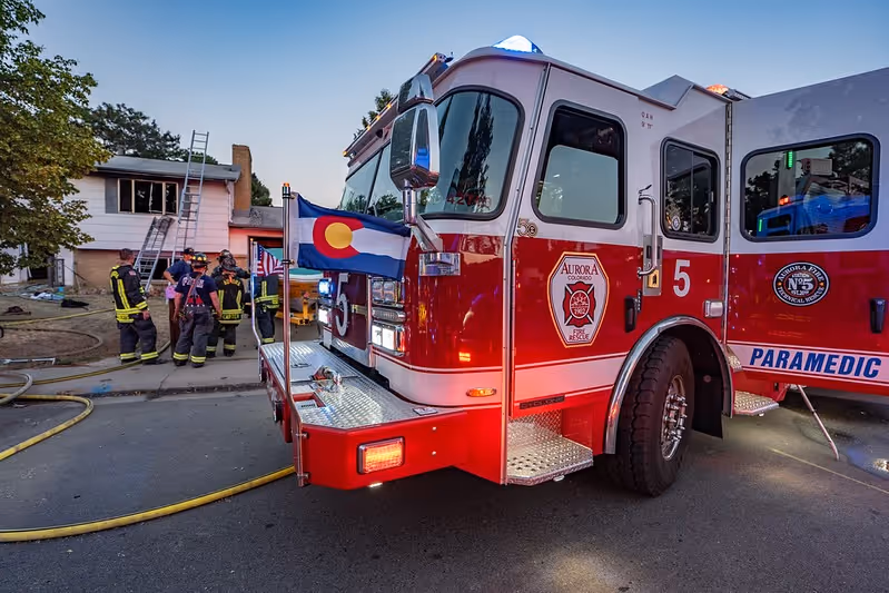 Red Aurora fire rescue truck parked near a house with firefighters and Colorado state flag during response.