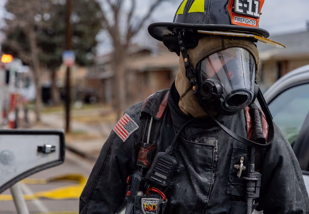 Firefighter in full gear and mask standing near emergency vehicles on a street.