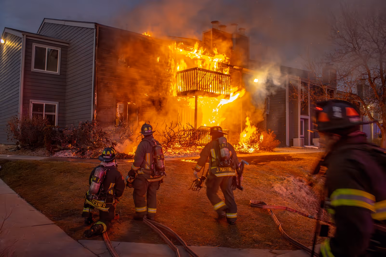 Firefighters in gear approach a blazing apartment balcony with flames and smoke at dusk.