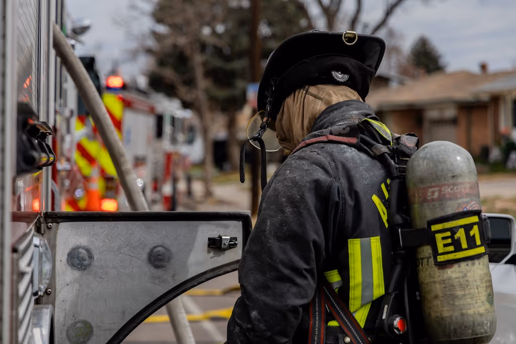 Firefighter in gear with air tank labeled E11 standing by fire truck with hose in background.