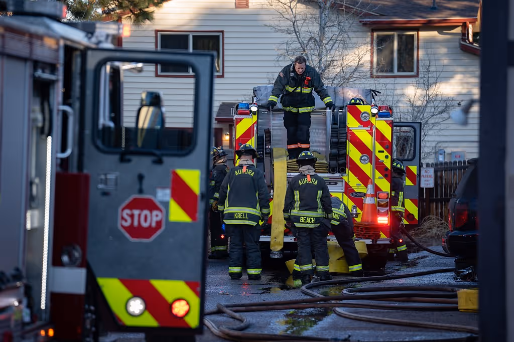 Firefighters in Aurora gear handling hoses at rear of fire truck near residential buildings.
