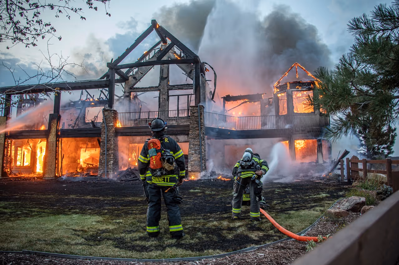 Two firefighters in gear spraying water on a large two-story house engulfed in flames.