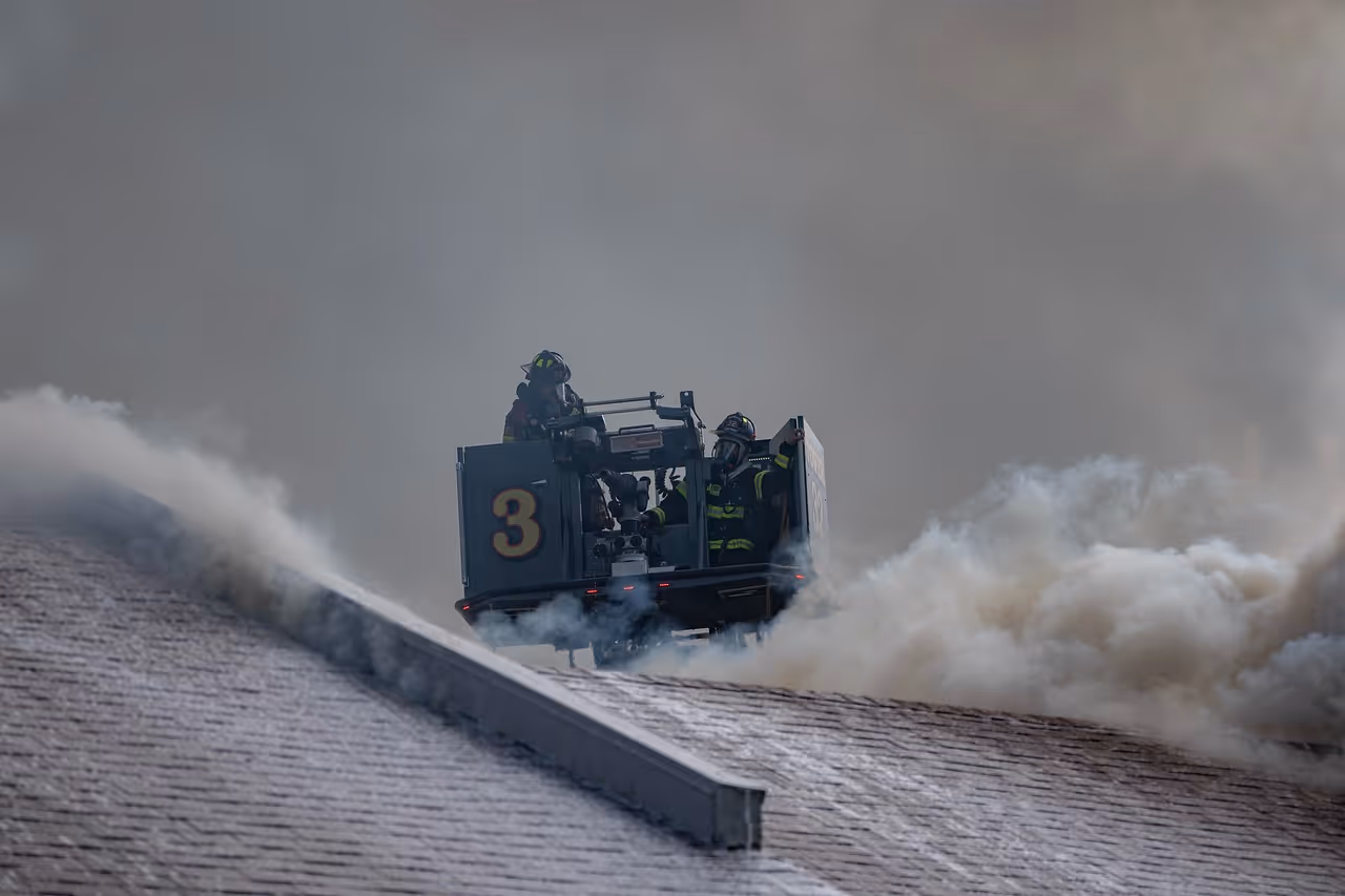 Firefighters in gear on a ladder truck spraying water on a smoky rooftop fire.