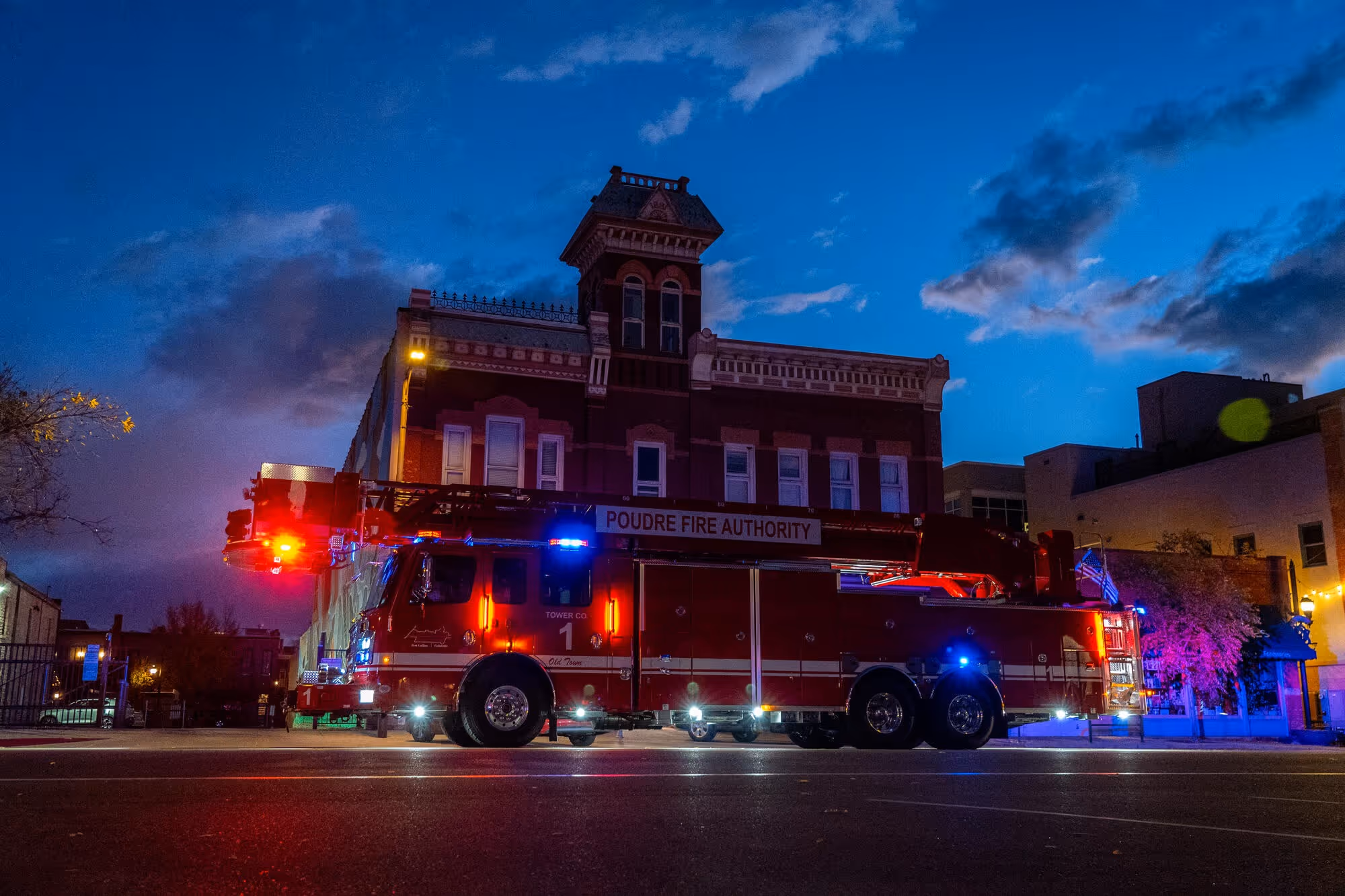 Red Poudre Fire Authority fire truck with lights on parked in front of a historic brick building at dusk.