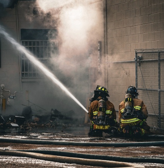 Two firefighters in full gear spraying water on a fire near a brick building and fence.