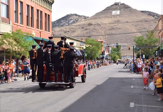 Group of firefighters riding a vintage fire truck in a parade on a town street with crowds watching.