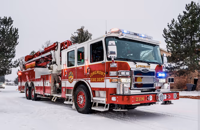 Longmont Fire Department red and white fire truck with lights on parked on snowy street.