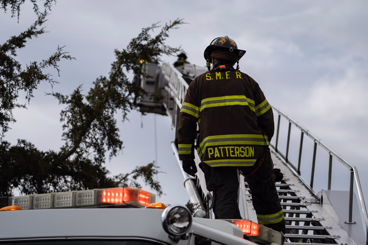 Firefighter with S.M.F.R. and Patterson on jacket climbs ladder on fire truck under gray sky.