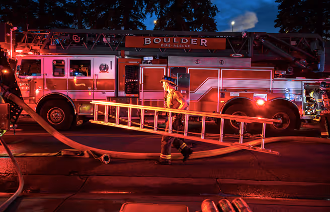 Firefighter in gear carrying a ladder past a Boulder fire rescue truck at night.