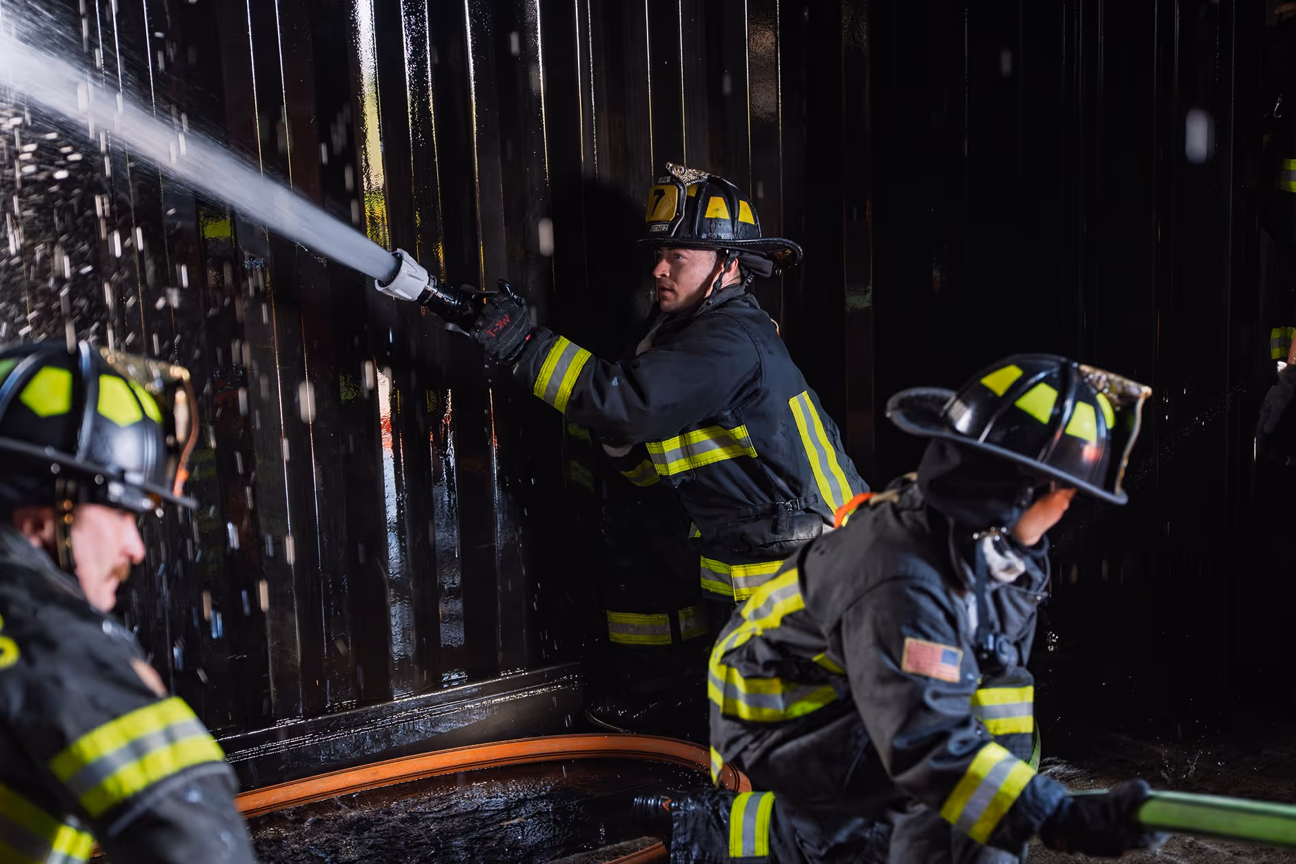 Firefighters in gear spraying water from hose inside a dark, wet area.