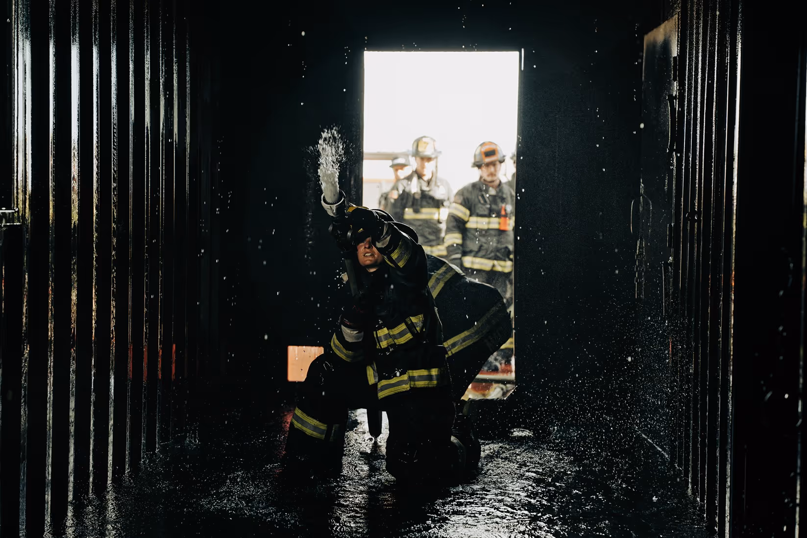 Firefighter kneeling and spraying water from a hose inside a dark, wet corridor.