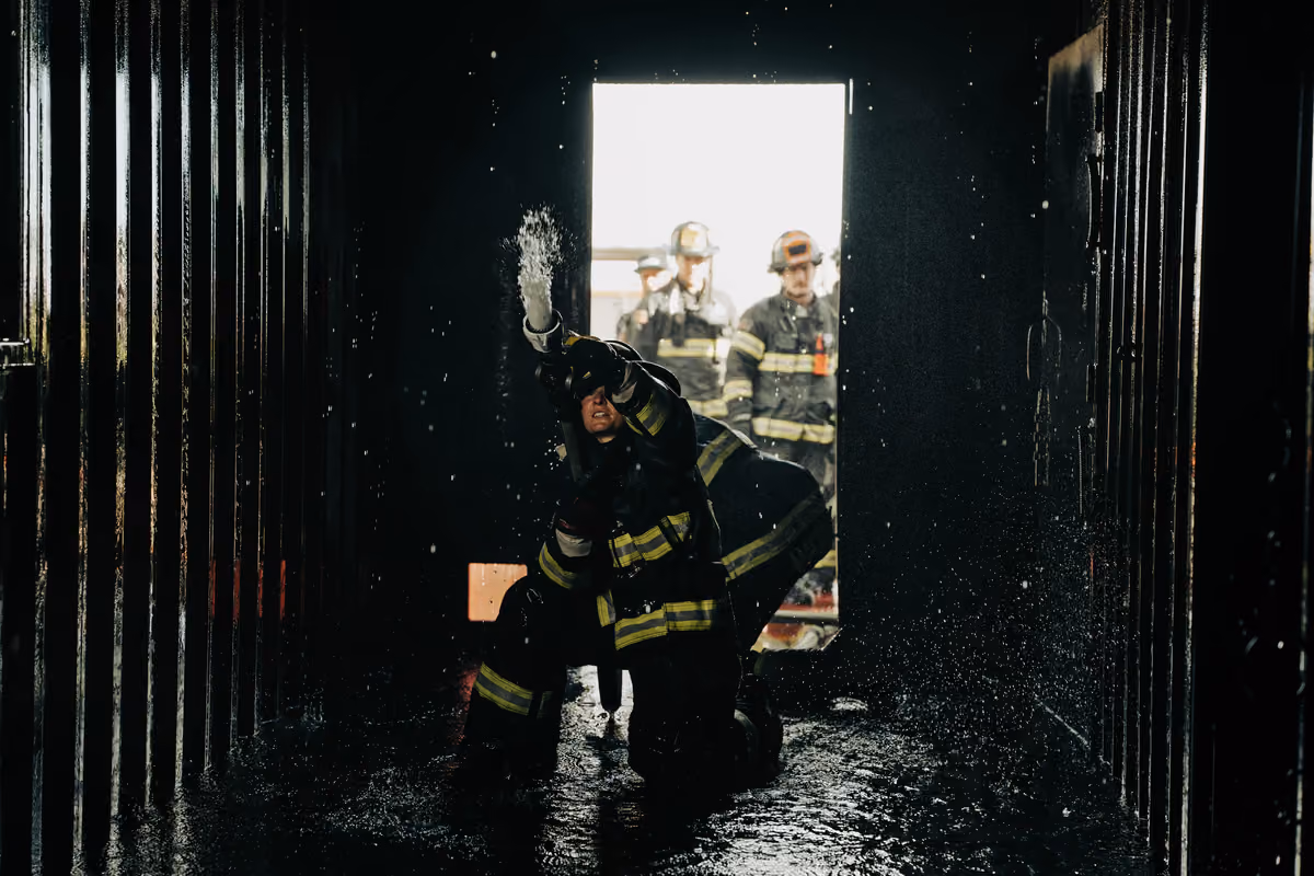 Firefighter in gear sprays water inside dark space while others watch from doorway.