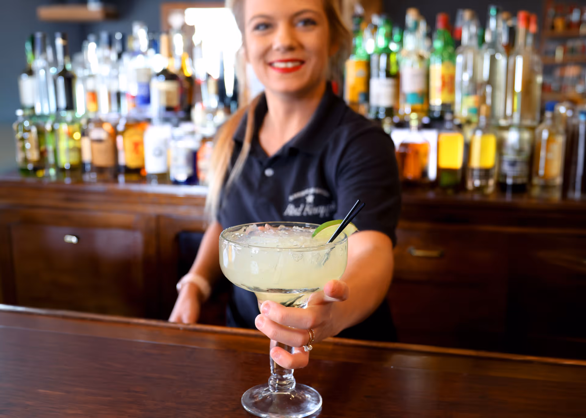 A bartender handing over a margarita glass with a lime wedge and black straw at a wooden bar.