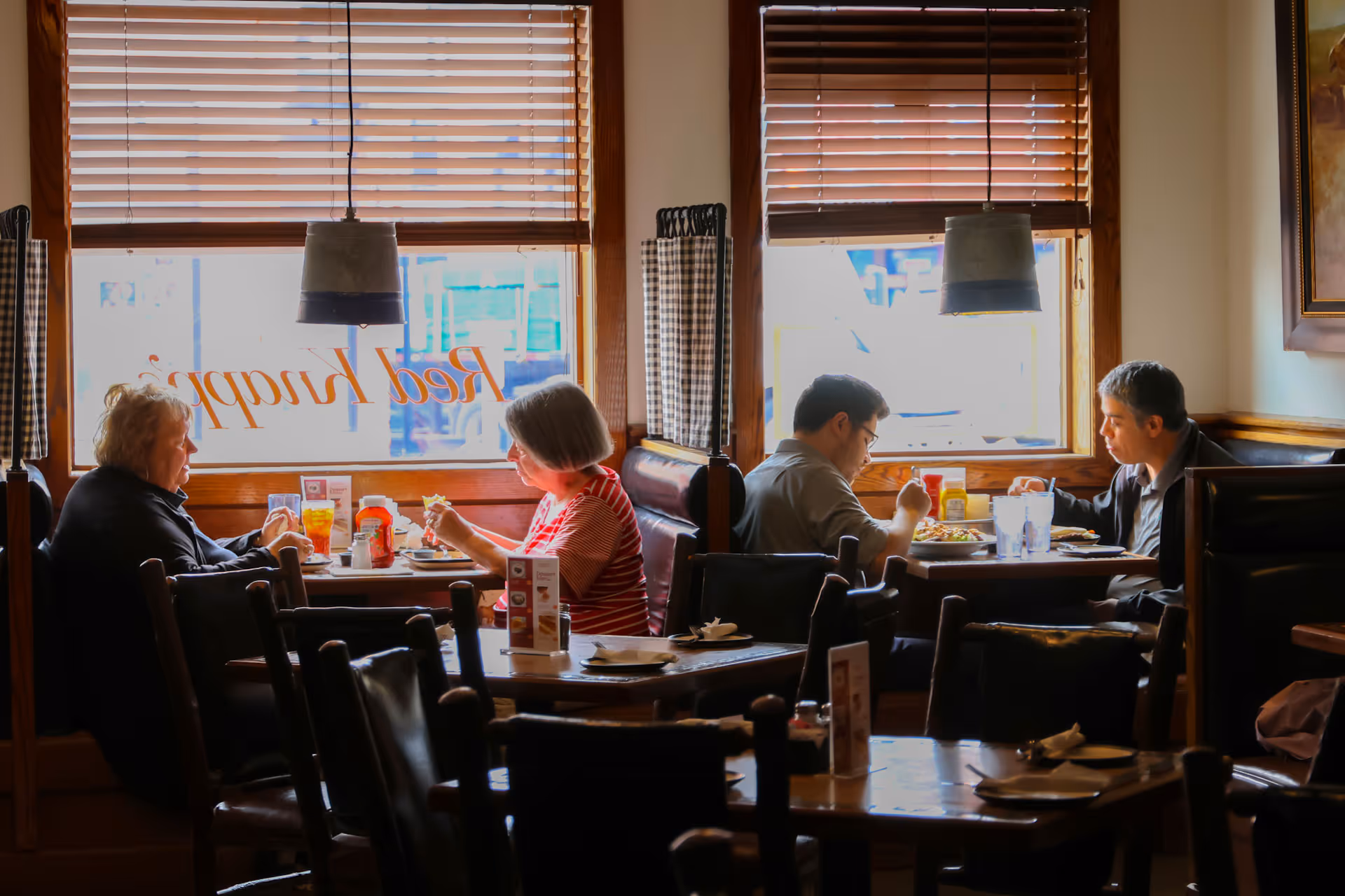 Four people sitting in pairs at two restaurant booths by windows with wooden blinds, eating and talking.