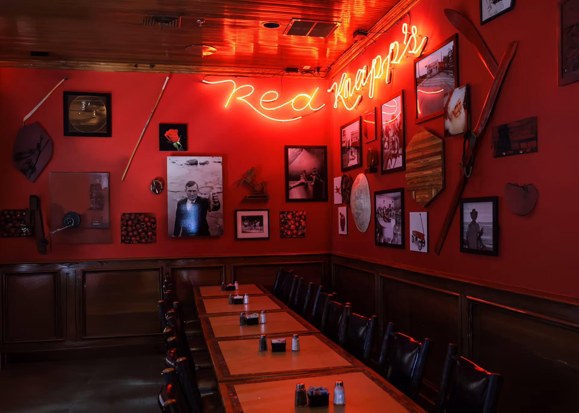 A long wooden table with black chairs in a red-walled room decorated with framed photos, vintage items, and a neon sign reading 'Red Knapp's'.