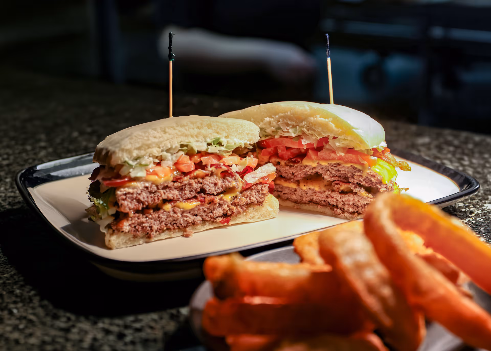 Double cheeseburger cut in half with lettuce, tomato, and pickles on a plate, with crispy fries in the foreground.