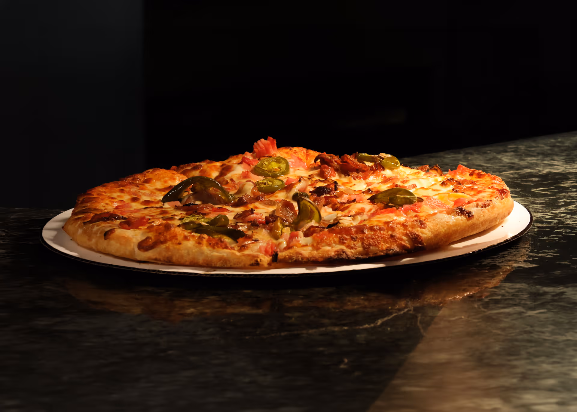 Hot pizza topped with cheese, jalapeños, and bacon on a white plate over a dark countertop.