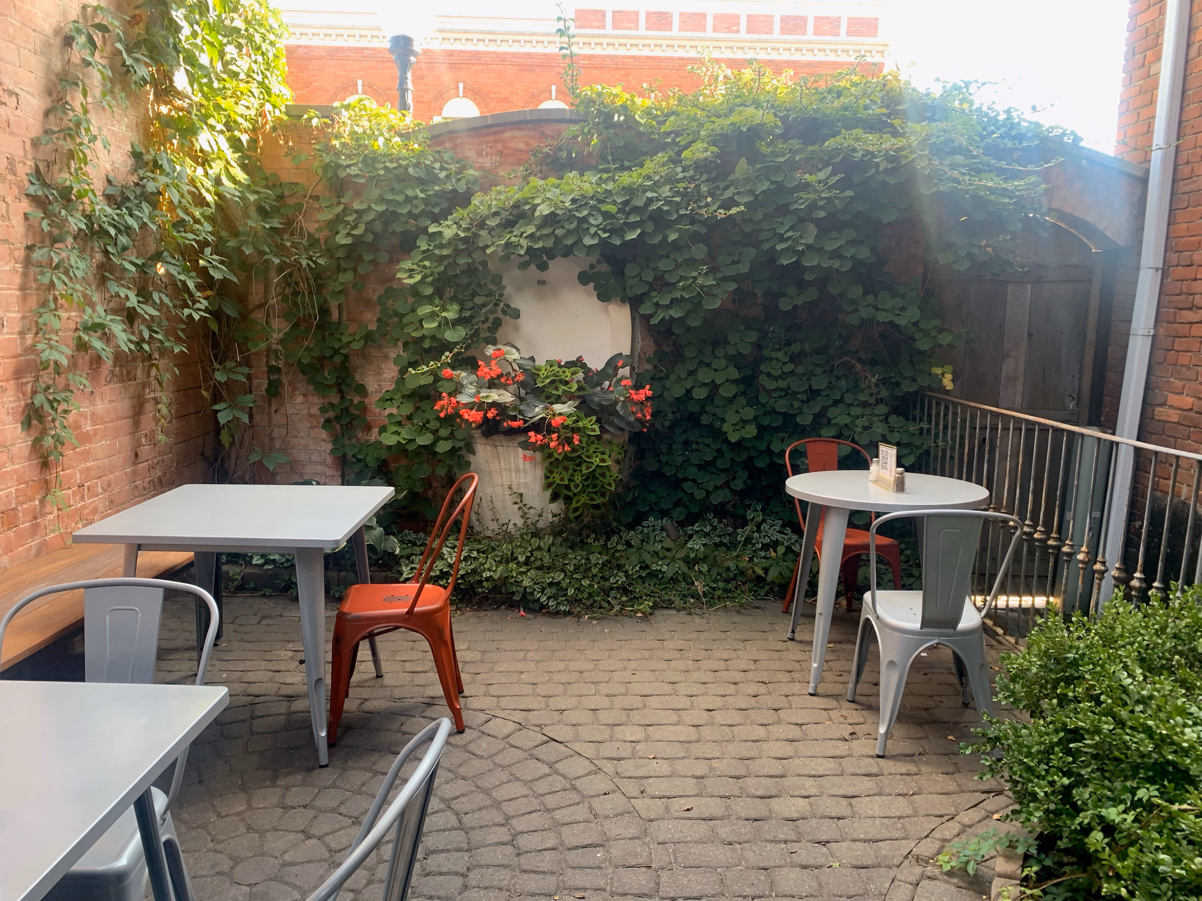 Outdoor patio with white tables, metal chairs, brick walls, and lush green vine-covered corner.