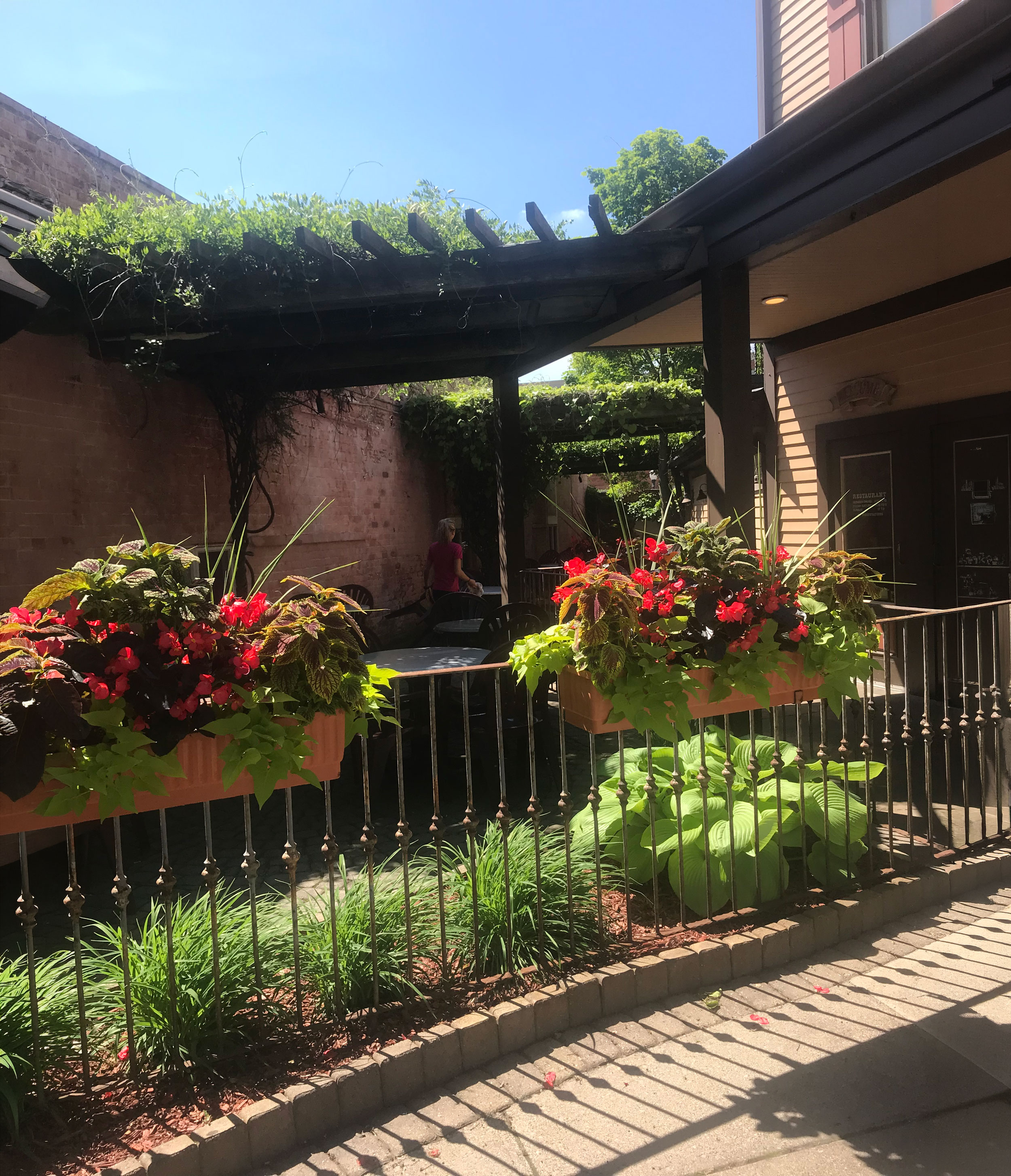Sunlit outdoor patio with hanging flower boxes, green plants, black railing, and a woman in background.