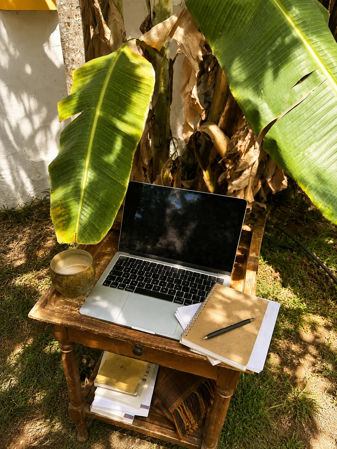Ordinateur portable et carnet posés sur une petite table en bois, en extérieur sous des feuilles de bananier