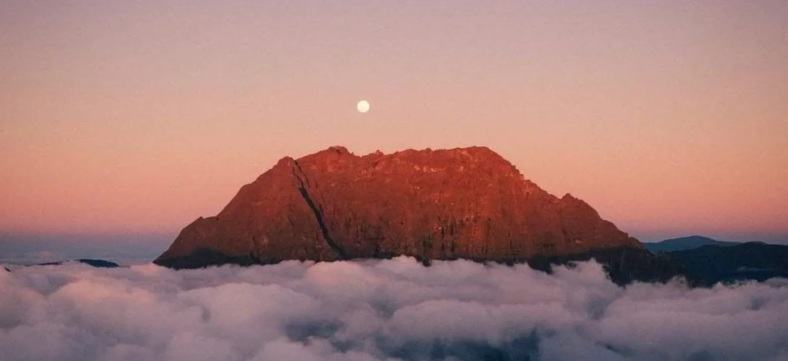 Sommet montagneux au-dessus d'une mer de nuages au coucher du soleil, avec la lune dans le ciel