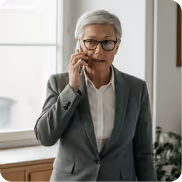 Older woman in gray suit and glasses talking on a smartphone indoors near a window.