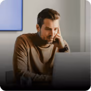 Man in brown sweater focused on laptop screen indoors with window in background.