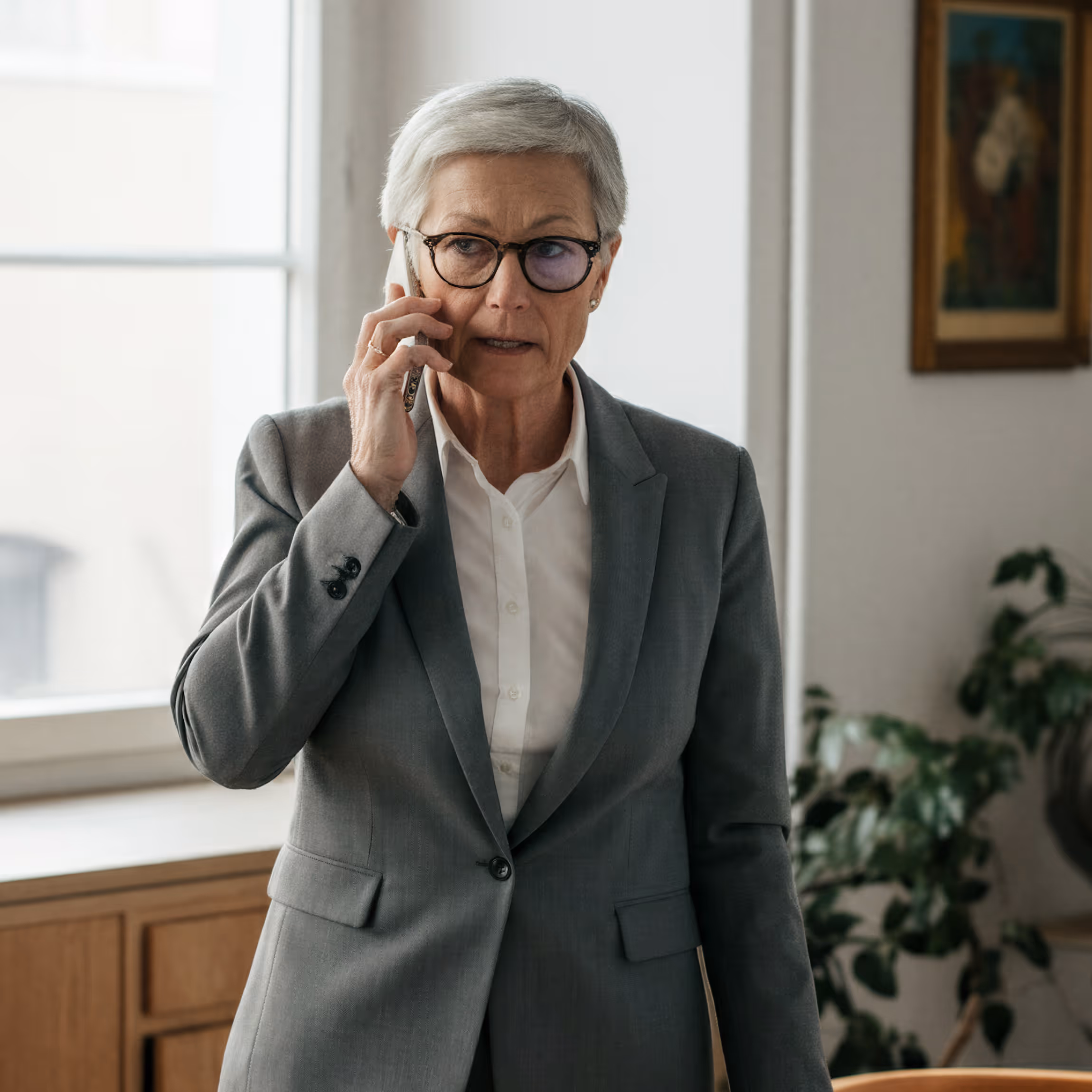 Mature woman in gray suit and glasses talking on a smartphone in an office setting.