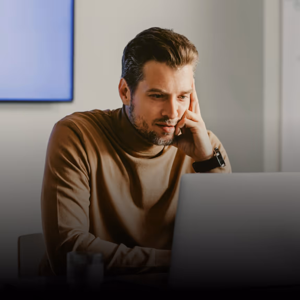 Man with a beard wearing a brown turtleneck and watch looking at a laptop screen in an office.