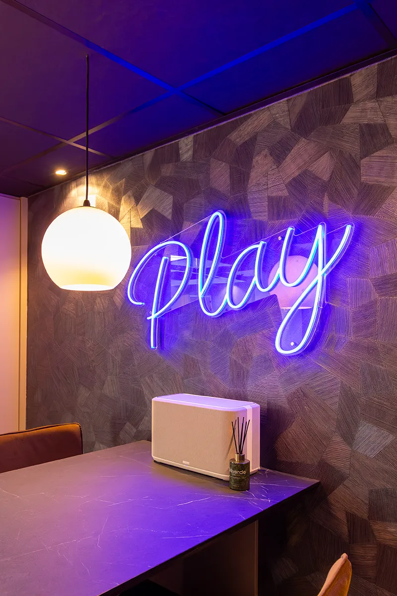 Detailopname van blauw neonbord met tekst Play en ronde hanglamp tegen donkere structuurwand in kantooromgeving