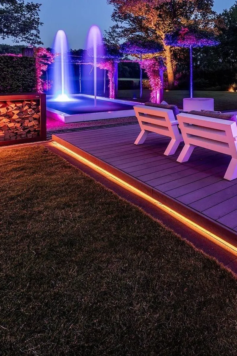 Luxe tuin in de avond met oranje LED-terrasrand, verlichte fontein en roze LED-boomverlichting op de achtergrond