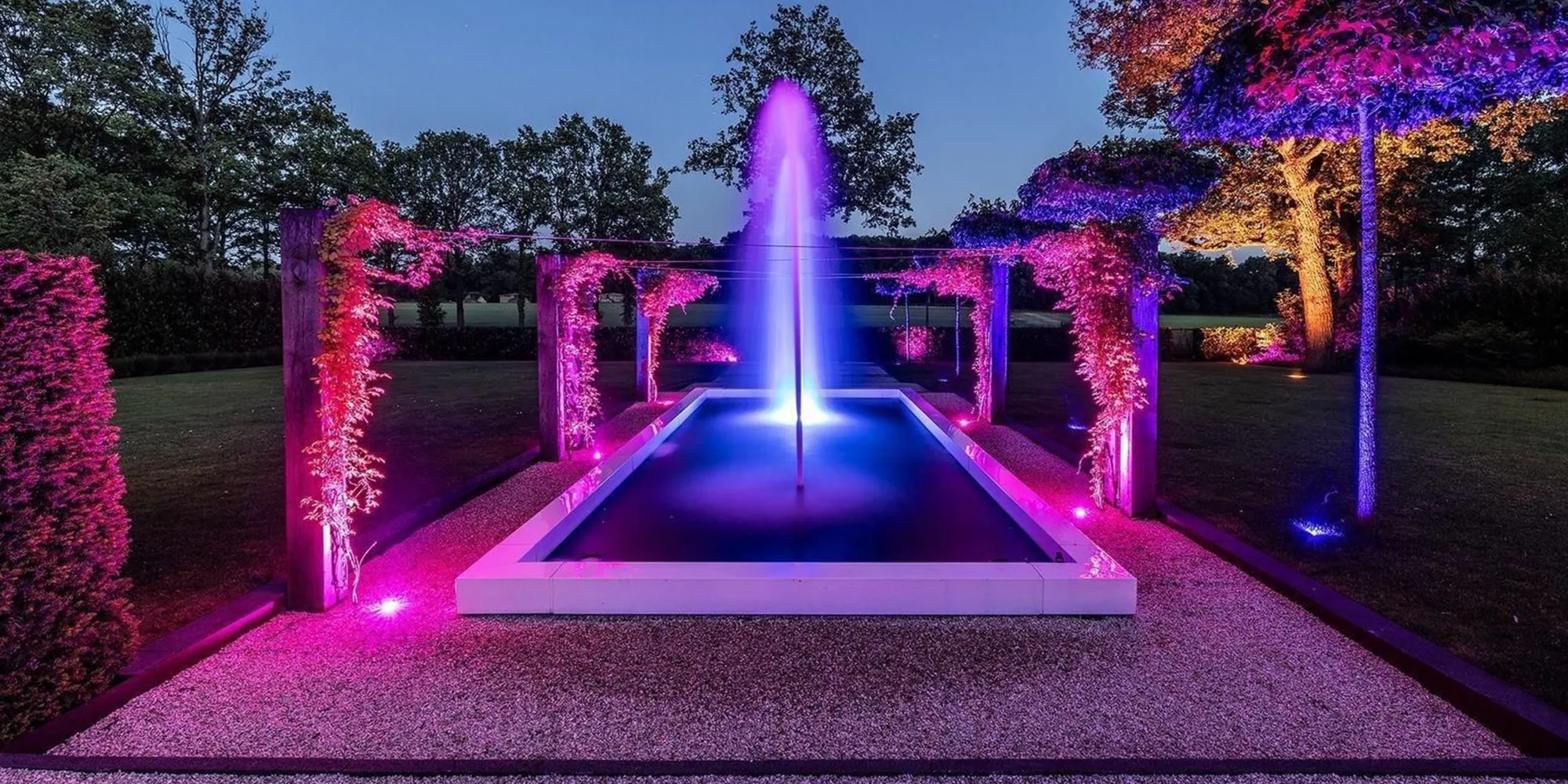 Breed overzicht van luxe tuin met verlichte fontein, roze LED-spots op klimplanten en bomen als compleet buitenlichtplan