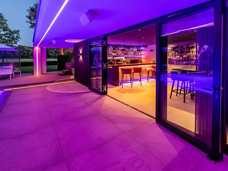 Overdekt terras van villa met paarse LED-verlichting en doorkijk naar warm verlichte huisbar met barkrukken