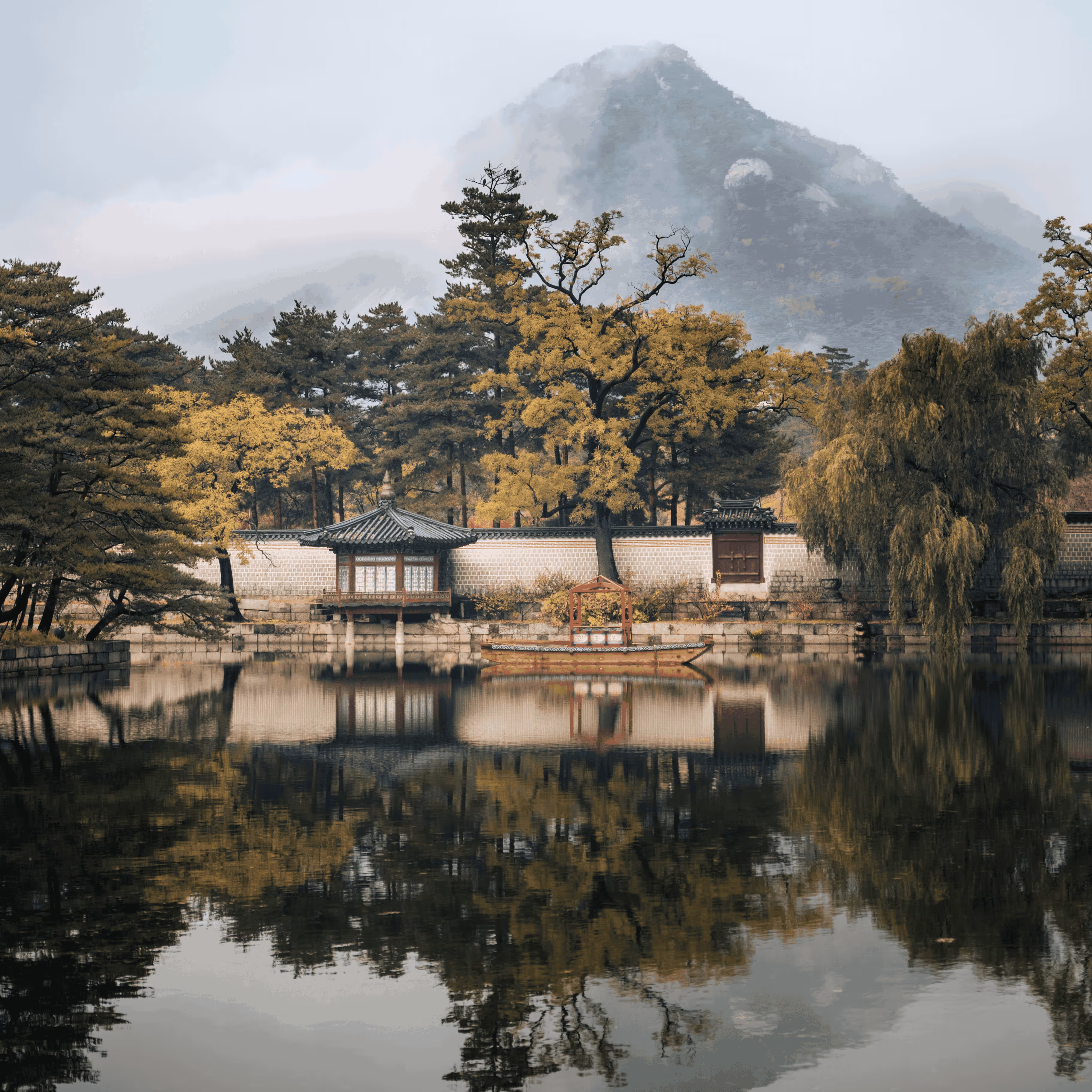 Traditional pavilion by a calm pond with autumn trees and misty mountain in background.