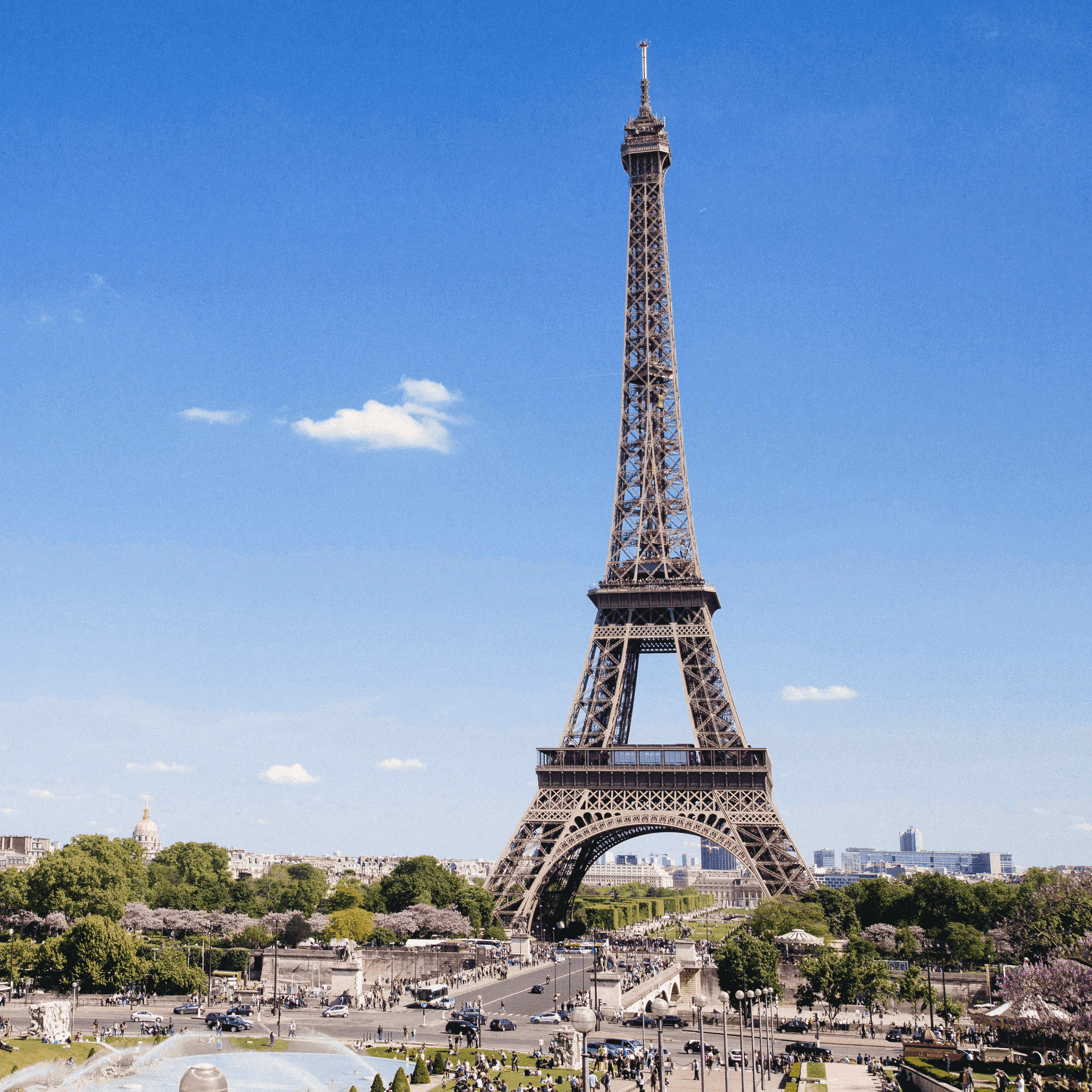 Eiffel Tower in Paris under blue sky with people, vehicles, and greenery in the foreground.