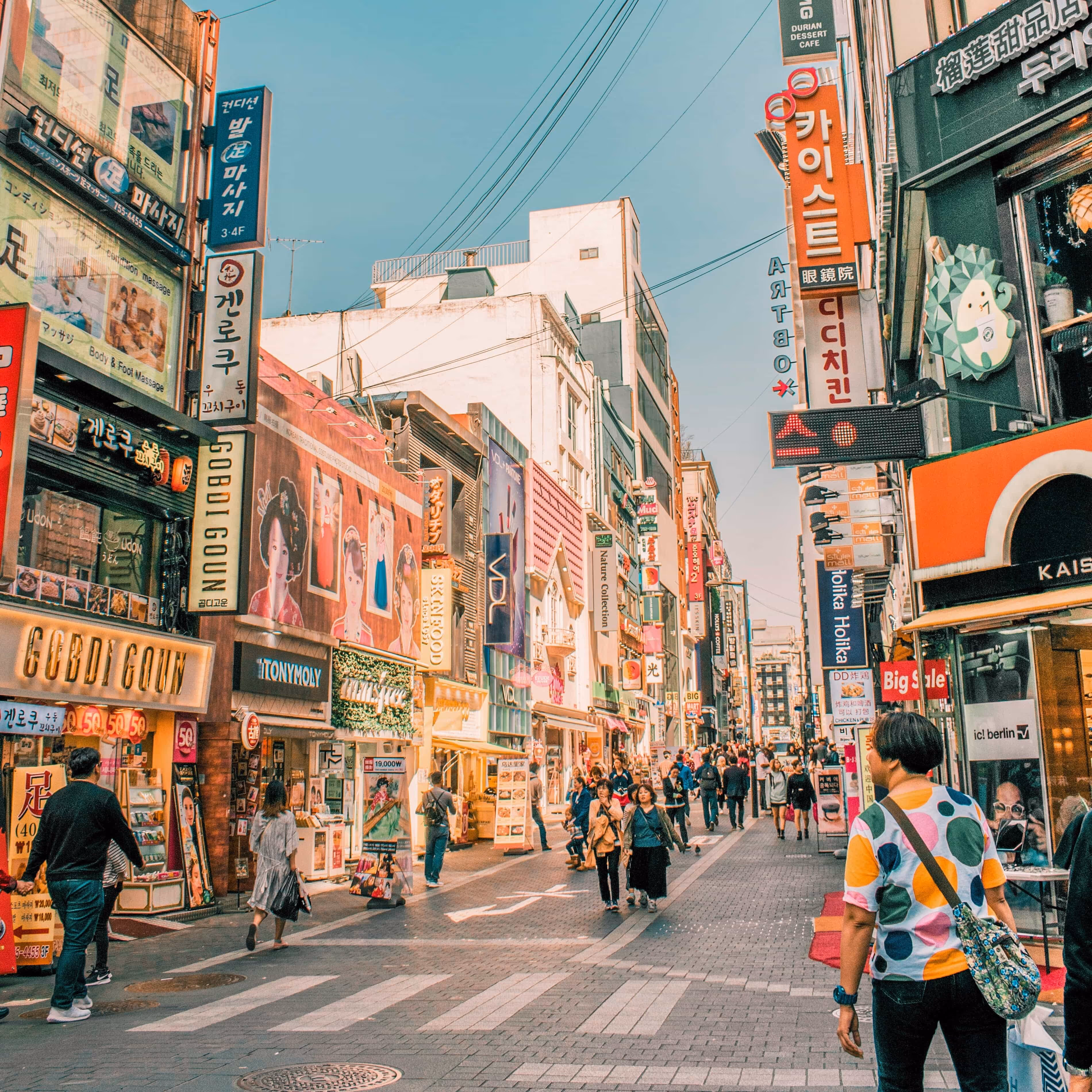 Crowded urban street with people walking past shops and colorful signs in Korean characters.