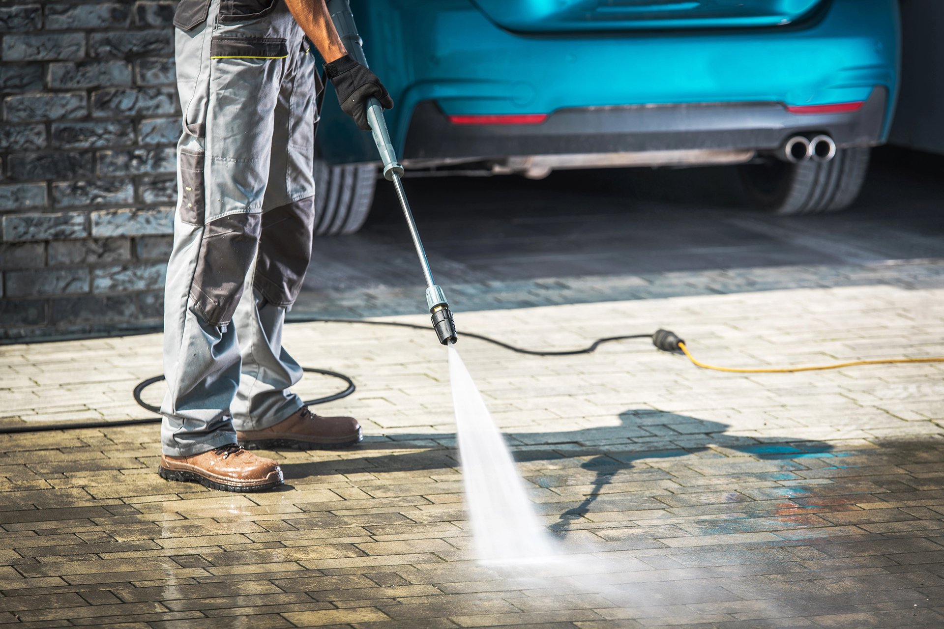 Person using a pressure washer to clean outdoor pavement near a blue car.