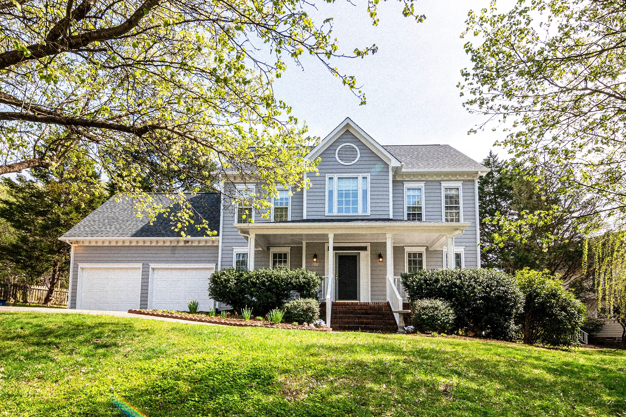 Two-story gray house with white trim, front porch, two-car garage, and green lawn with trees.