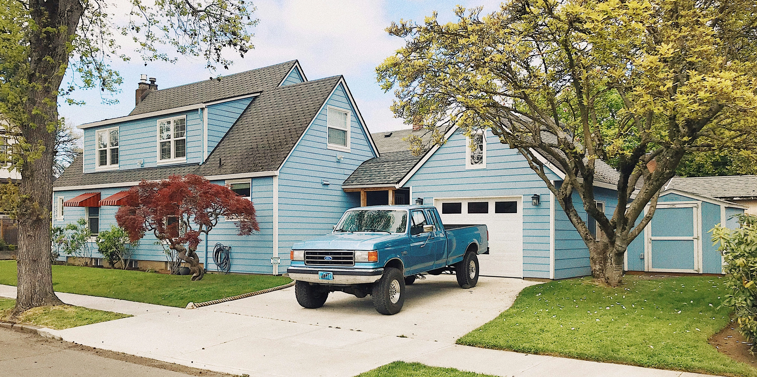 Two-story gray house with white trim, front porch, two-car garage, and green lawn with trees.