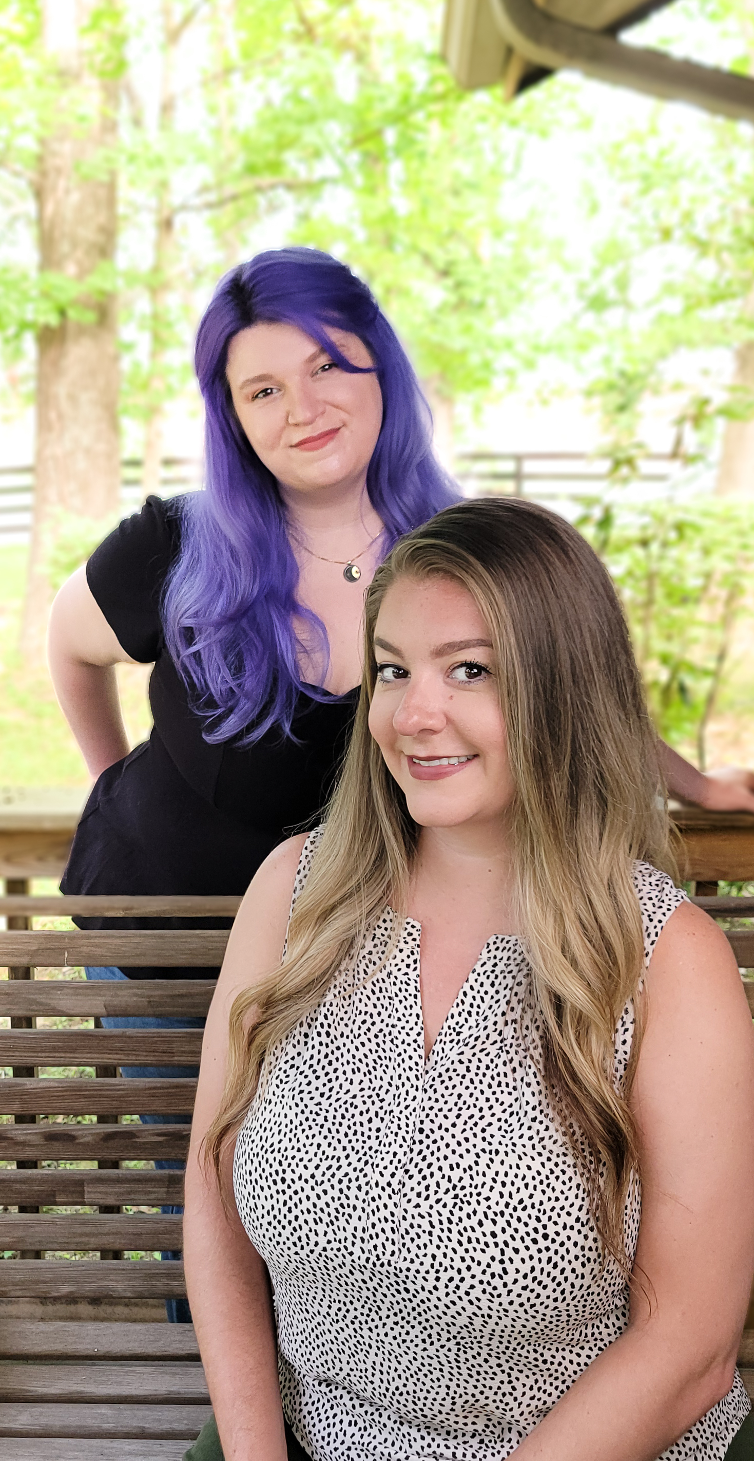 Amanda Hargrove and Katie Sanchez smiling outdoors on a wooden bench; one with purple hair, Amanda, stands behind the blonde seated woman, Katie.
