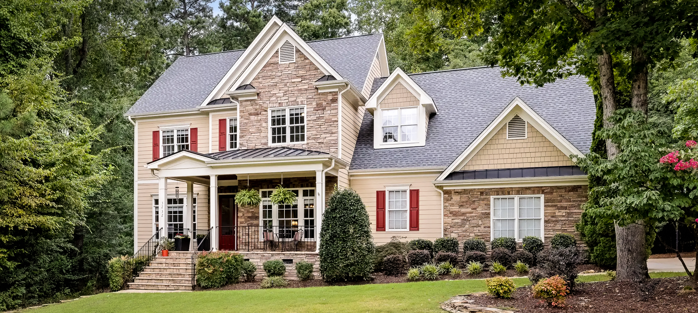 Two-story gray house with white trim, front porch, two-car garage, and green lawn with trees.