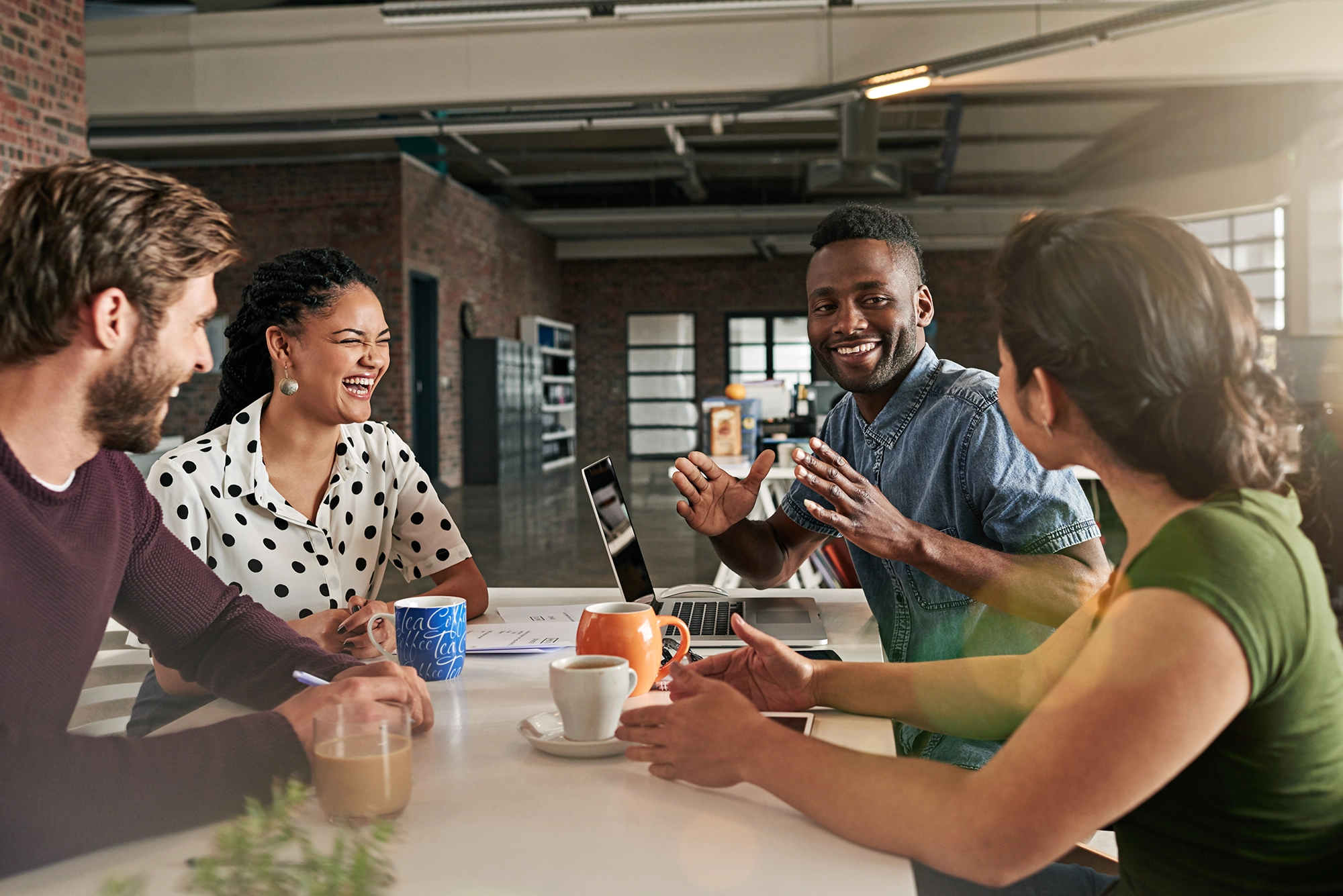 Four coworkers in a casual office setting smiling and having a lively discussion around a table with laptops and coffee cups.