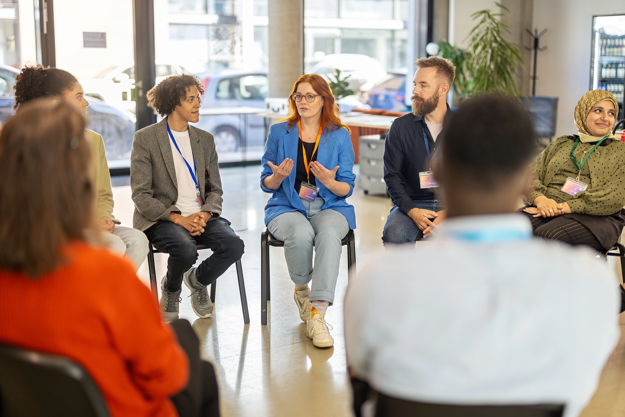 A diverse group of people seated in a circle in a bright room, engaged in a discussion with a woman in a blue blazer speaking.