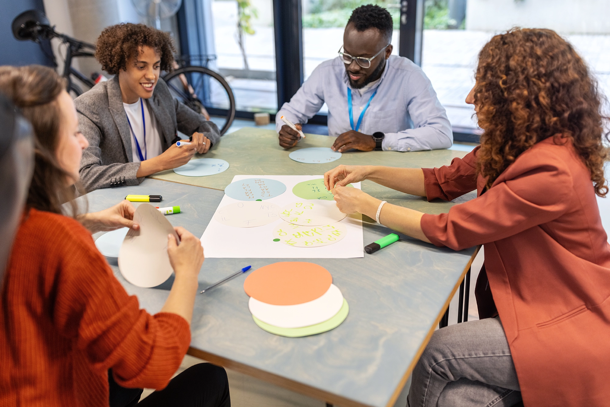 Four people sitting around a table in a meeting, writing on round colored paper cutouts and arranging them on a larger sheet.